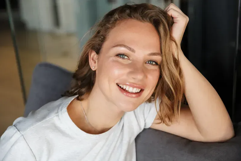 Smiling woman with wavy hair resting her head on her hand, sitting on a couch.