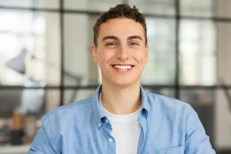 Smiling young man with curly hair wearing a light blue shirt in an office.