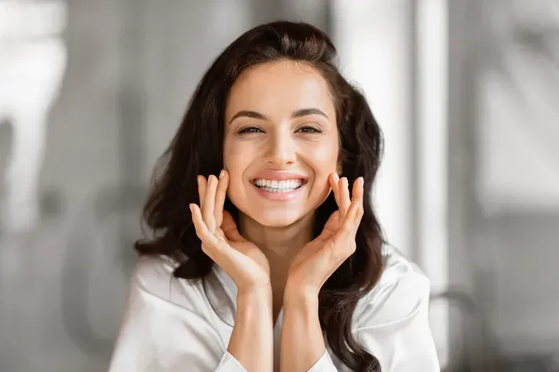 Smiling woman with dark hair touching her cheeks, wearing a white robe.
